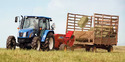 Blue tractor in hay field pulling red bale thrower that is ejecting rectangular bales into a brown hay wagon with 3 high sides. Blue sky background.