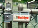 The deviceâ€™s components shown secured at the upper corner of a metal chain-link gate and on the fence post to which is also affixed the push-button control pad.