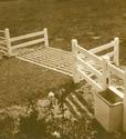 The white, slotted cattle guard lying flush on the soil surface with a white two-rail fence section on each side and connected to the coralâ€™s three-rail fencing.