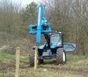The post-driver unit front-mounted on a teleloader with the steel tube above one of the two round wooden posts that has just been driven into the ground.