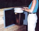 An adult female fills a feed bucket bolted to the inside of the door thatâ€™s hinged to metal-framed 2 x 2-foot opening in the front of a pen.