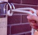 Manâ€™s hand holding a thick rope thatâ€™s coiled around this deviceâ€™s the shiny metal rings which extend out from a base plate thatâ€™s bolted to a vertical 2x4.