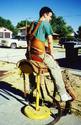 Astride a saddle is an adult male wearing the harness with the wide chest/abdomen belt seen anchored to the back of the saddle and the over-the-shoulder straps that clip in front to the saddle loops.