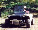 Front view of the vehicle being operated by an adult male; seen is the forward electric winch, the windshield, and the ROPS.