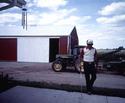 An adult male (with cane) walking from barn toward house, which has a wind-chime set hanging from the corner eave.