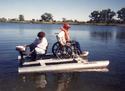 Two adult males aboard the boat, one toward the front seated in his wheelchair, the other in the back operating a trolling motor.