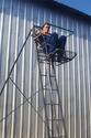 An adult male seated in the already elevated hunterâ€™s compartment of a tall ladder stand thatâ€™s leaning against a building.