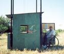 Two photosâ€”(1) the blind elevated to the top of the standâ€™s four steel-tube frame; (2) the blind at ground level, its door open and an adult male in a wheelchair entering.