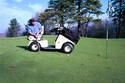 Putting on a golf course green is a lower-extremity adult male amputee on the swivel seat of a Solorider, his club-bag anchored onto the cartâ€™s hood.