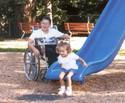 Boy in a wheelchair and little girl coming off a slide in a play area that has a woodchip surface.