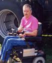 An  adult male in a power wheelchair holding the CB mic, its cord running to the transceiver on the flat fender above the chairâ€™s left drive wheel.