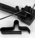 The vice-mounted tool in the process of bending a portion of round stock, and a piece of flat stock that had already been bent as the user desired.