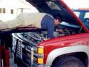 With the creeper frame in front of the truck and the hood up, an adult male, lying face down on the deviceâ€™s horizontal platform, is reaching down into the engine making a repair.