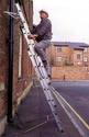 An adult male climbing a three-unit extension ladder. with the safety attachment affixed to the bottom ladder second rung.