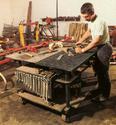 Adult male cutting a piece of metal over the grill portion of his portable welding table, with its cater wheels, pipe legs, hanging wrenches and clamps, plus water bucket clearly visible.