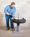 With it resting thigh-high and horizontally on the changer, an adult male is removing tire from wheel using the mount/dismount bar.