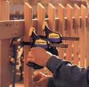 Making a wooden fence, an adult male squeezes the device pistol-grip trigger to clamp a 1x2 vertical picket against the 2x6 horizontal board, ready for nailing.