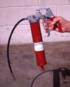 Affixed onto the top of the grease-filled cylinder is a unit consisting of the delivery hose and a pistol-grip pumping mechanism that an adult male hand is squeezing.