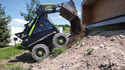 Black and green 4-wheeled skid steer dumping dirt from its front-end bucket on an up-hill dirt approach to a brown wood building. Green grass-white building-port-a-pot and blue skies with white clouds in background