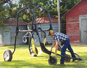 Man on rt in blue jeans checked shirt and cap bending over pushing large tubular fram on 4 wheels with 1 metal sling & 1 strap sling suspended from pulleys within the frame.Red barn & 2 round metal bins in background