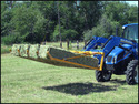 Blue New Holland tractor with yellow bale sweep that looks like a 5-tine fork carrying 4 rows of small square hay bales in a green pasture with green trees