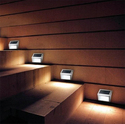 Brown stairs in front against a brown horizontally-lined wall with a silver solar light box attached to the wall for each stair-step illuminating the step and the riser to the next step.