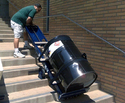 Man in green t-shirt and white shorts guiding a blue 2-wheeled powered dolly with a black 55 gal metal barrel on it up cement steps on the outside of a brick building