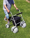 Person in short sleeve shirt & calf-length blue jeans pushing silver tubular frame walker with large baloon-type tires on green grass