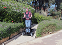 Man wearing red & gray checked shirt & blue jeans in standing position in upright wheelchair on sidewalk between flowering hedges and gardens
