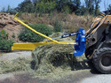Blue and yellow round-bale slicer on front end of yellow skid steer slicing a bale of hay with a hill & green bushes in background