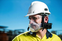 Man from shoulders-up wearing yellow & black shirt - white hard-hat - goggles - & a clear respirator face mask with a battery pack around the back of  his neck. Blue sky in background.