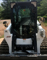 Black & white skid steer with round side-view mirrors mounted on each side of cab on skid steer body. Dirt road & pine trees in background.