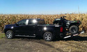 Black pickup truck with ATV half in pickup bed & half on ramp that is sloping upward from the fender holding the ATV in place. Brown corn field in background.