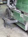 Green forklift with horizontal metal rod bolted onto lower side panel at wheel axle height and metal step fastened to metal rod with chain to pull it up or let it down and metal stand welded to step for the down position.