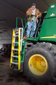 Man in blue jeans brown coat and black cap standing on top of platform on large green farm equipment & holding silver chain attached to yellow step ladder that is fastened on outside of standard green steps integral to the farm equipment