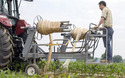 Red tractor pulling gray platform on wheels with man standing on back of platform. Platform has 2 automated spindles lifting and rolling up plastic sheets from the field.