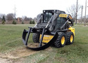 Black and yellow skid steer in yard with an attachment looking like a half-circle with two arms attaching one third of the way in from the sides. Attachment can be raised or lowered with 2 hydraulic pistons.