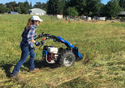 Woman in blue jeans checkered shirt & white baseball cap guiding a walk-behind tractor in a grassy field with farm pens and metal shed in background.