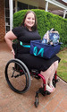 Smiling woman in black dress sitting in wheelchair with dark blue cloth box with light blue cloth handles on her lap & a backpack & water bottle in the box.