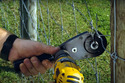 Man's hand holding black wire-twister tool with yellow drill attached to it against a wire fence with grass in the background