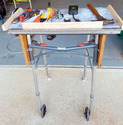Standard metal walker frame with 2 small wheels on front legs and feet on back legs with aluminum table top mounted on top of the walker & wood pieces fastened on the top edges to keep tools from falling off