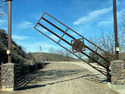 4-horizontal-bars metal gate sloping from post on right up toward the left in raised position above a gravel road & against a blue sky