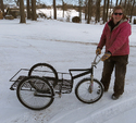 Woman in pink winter coat & brown snow pants on right holding handlebars of 3-wheeled low bicycle-tired metal cart on snowy ground with woods in background