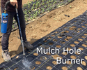 A person from the waist down holding a mulch hole burner over a row of black plastic marked off in squares in a garden