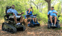 2 men and a woman each sitting in an EcoRover Tracked Wheelchair in a woods with a dog standing next to the man on the left and the woman in the center holding binoculars.