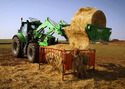 Green tractor in field with Unrolla Round-Bale Unroller mounted on it dispensing hay into feeding bin