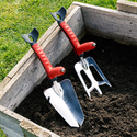 2 red-handled Multi-Digger Hand Tools sitting in some dirt in a raised bed garden.