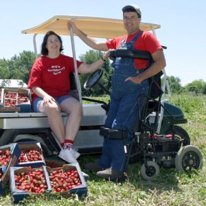 Ed Bell using standing wheelcair in front of golf cart filled with strawberries with wife sitting on cart