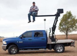 Photo of man sitting on lift attached to bed of pickup truck. He is about 10 feet off the ground.