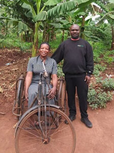 Mutumba Faisal stands next to woman sitting in hand-powered, three-wheeled bicycle used for mobility with tropical foliage in background.